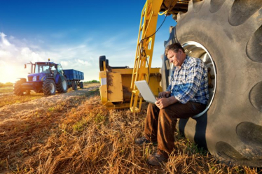 Farmer in a field with a laptop and a tractor in the background.